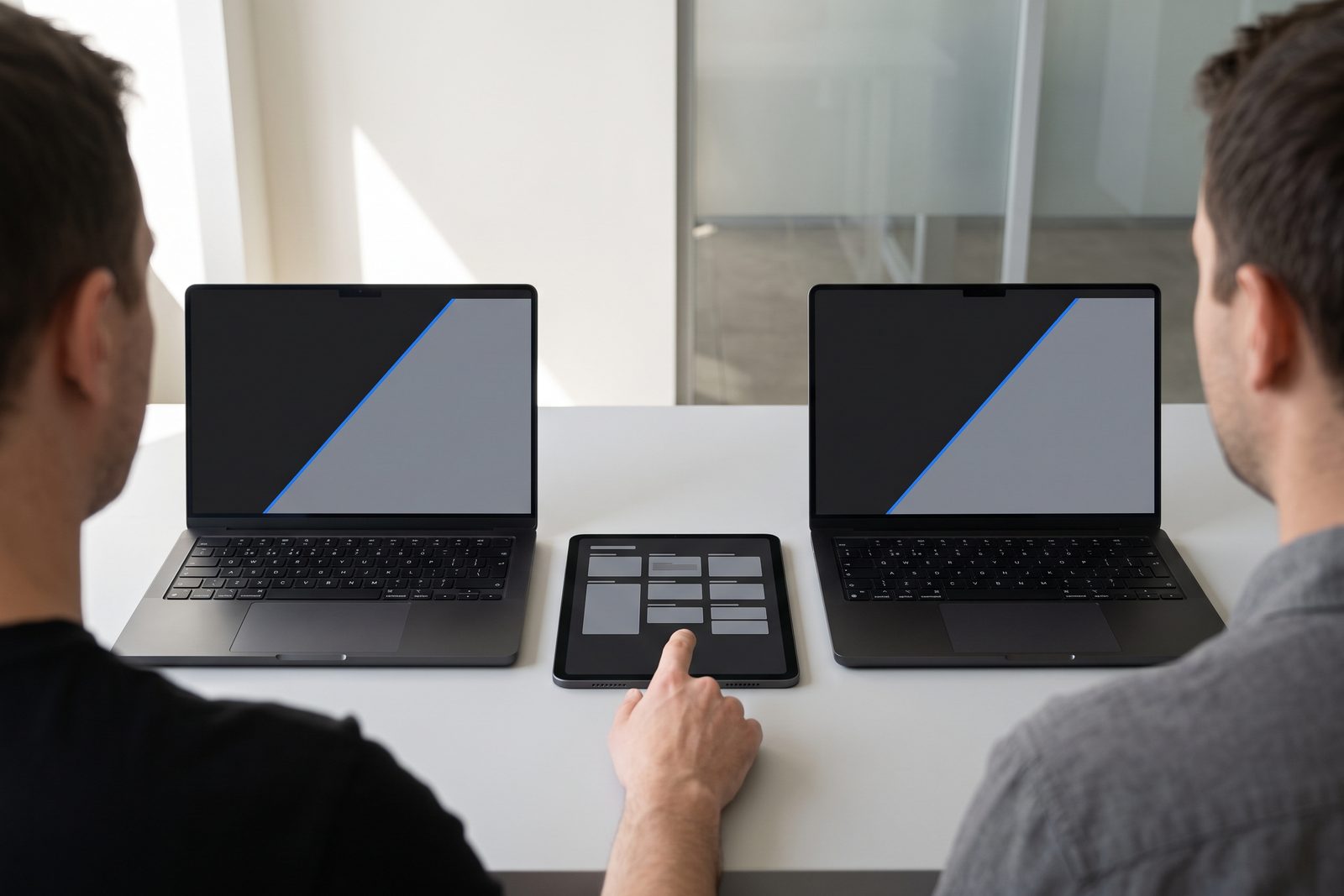 Two engineers at a handover desk with two Space Black MacBook Pros and an iPad Pro showing the runbook